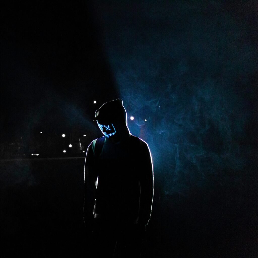 Silhouette of a man in a glowing LED mask and hoodie in a dark, foggy setting.