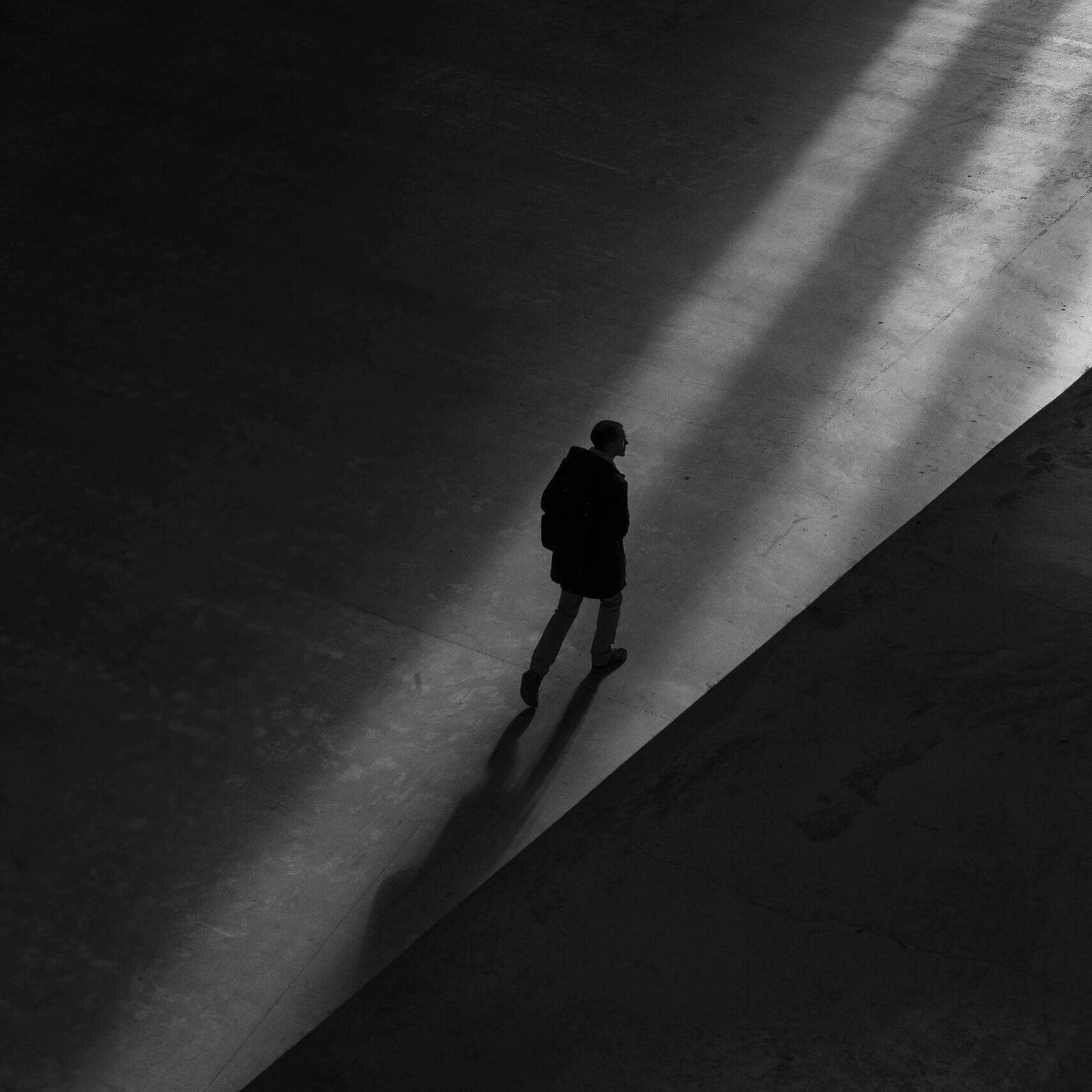Black and white photo of a man walking through shadows on a deserted pavement, creating a moody silhouette.